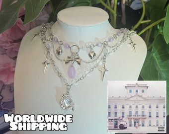 May include: A layered silver necklace with purple and clear beads, heart charms, and star charms. The necklace is on a white mannequin. The background is a blurry image of a house with the text "B-12" on the side.