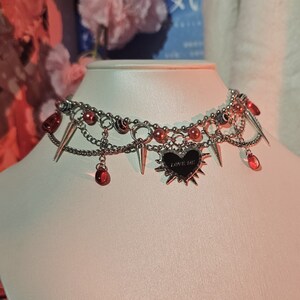 May include: A silver choker necklace with multiple chains, red beads, and spikes. A black heart charm with the text "LOVE ME" hangs from the center.