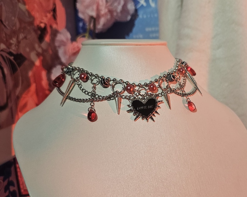 May include: A silver choker necklace with multiple chains, red beads, and spikes. A black heart charm with the text "LOVE ME" hangs from the center.