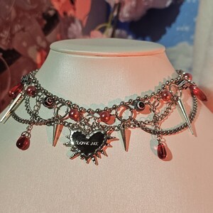 May include: A silver choker necklace with multiple chains, red beads, and silver spikes. The necklace features a silver heart charm with the words "LOVE ME" inscribed on it.