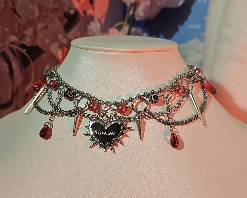 May include: A silver choker necklace with multiple chains, red beads, and silver spikes. The necklace features a silver heart charm with the words "LOVE ME" inscribed on it.