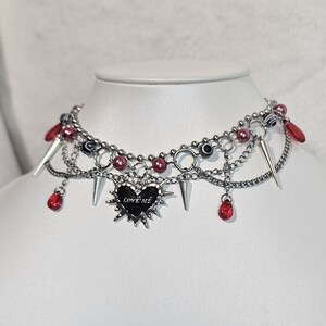 May include: A silver choker necklace with multiple chains, red beads, and a silver heart pendant with the words "LOVE ME" on it. The pendant is surrounded by spikes.