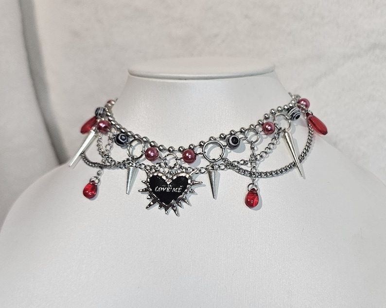 May include: A silver choker necklace with multiple chains, red beads, and a silver heart pendant with the words "LOVE ME" on it. The pendant is surrounded by spikes.