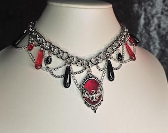 May include: A silver-toned choker necklace with red and black accents. The necklace features a chainmail design with dangling red and black teardrop-shaped beads. A red oval pendant with a bat design hangs in the center.