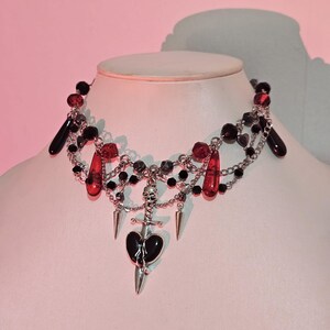 May include: A black and red beaded choker necklace with a silver pendant featuring a skull, a dagger, and a broken heart.