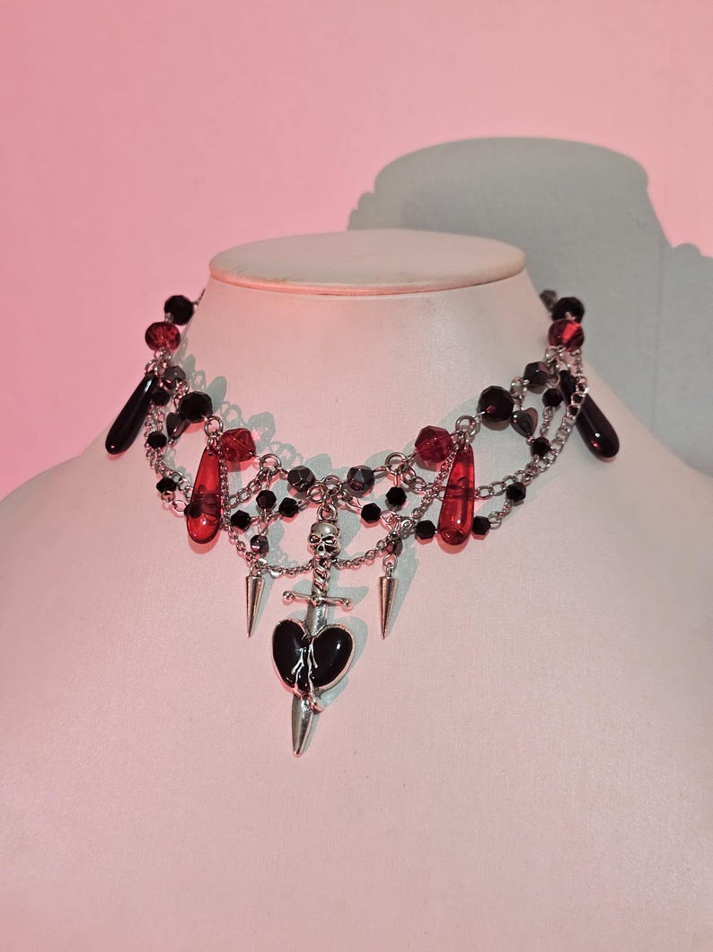 May include: A black and red beaded choker necklace with a silver pendant featuring a skull, a dagger, and a broken heart.