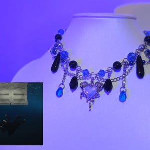May include: A silver chain necklace with a blue and black beaded design. The necklace features a dangling heart charm with a white center and a silver design. The necklace is on a white surface.