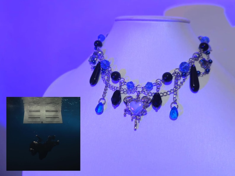 May include: A silver chain necklace with a blue and black beaded design. The necklace features a dangling heart charm with a white center and a silver design. The necklace is on a white surface.