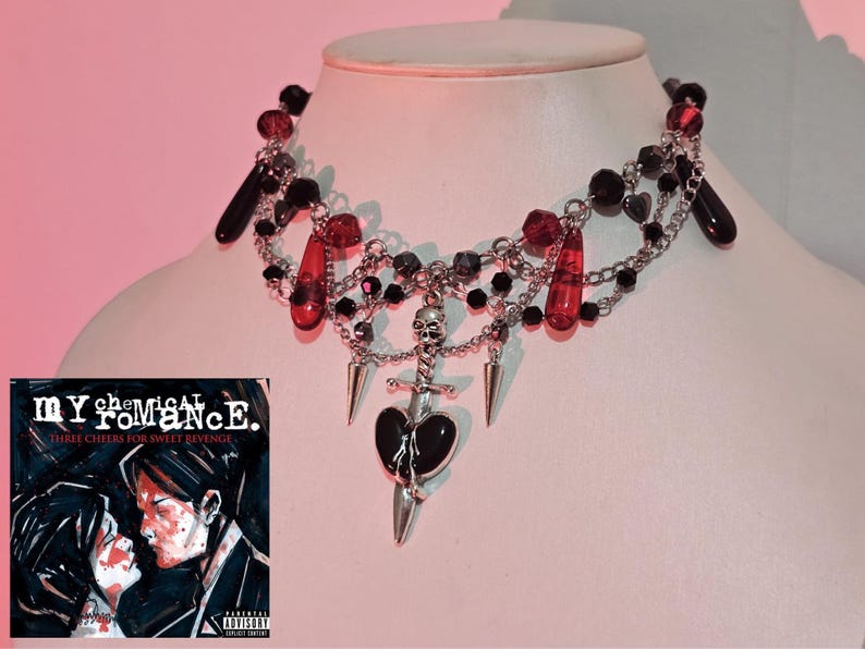 May include: A silver necklace with a black heart, a skull, and a dagger pendant. The necklace is made with black and red beads and silver chains. The necklace is on a white background. The album cover for My Chemical Romance's "Three Cheers for Sweet Revenge" is in the background.