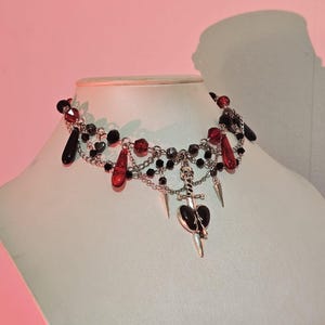 May include: A silver choker necklace with black and red beads, a silver skull charm, and a silver dagger charm with a black heart.