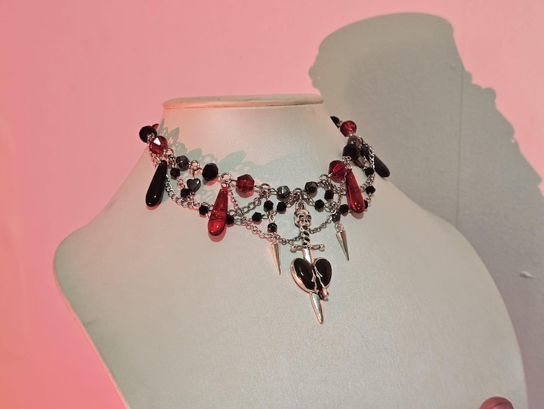 May include: A silver choker necklace with black and red beads, a silver skull charm, and a silver dagger charm with a black heart.