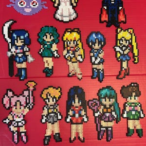 Könnte beinhalten: Eine Sammlung von 12 Pixel-Art-Charakteren aus der Anime-Serie Sailor Moon. Die Figuren bestehen aus bunten Plastikperlen und beinhalten Sailor Moon, Sailor Mercury, Sailor Mars, Sailor Jupiter, Sailor Venus, Tuxedo Mask und Luna.
