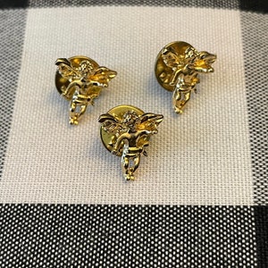 May include: Three gold-tone cherub pins with wings and a halo. The cherubs are holding a heart in their hands.