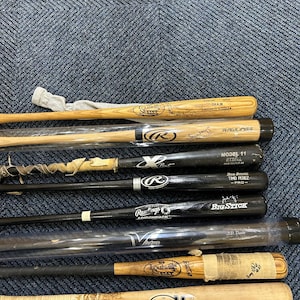 May include: A collection of baseball bats, some with signatures and brand names like Rawlings and Louisville Slugger. The bats vary in color, with some being natural wood and others black. Some bats are wrapped in plastic or tape.