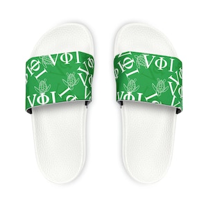 Pode incluir: Sandálias brancas com uma tira verde com letras gregas brancas e ilustrações de tartarugas. As sandálias têm uma palmilha branca texturizada e um forro interior preto na tira.