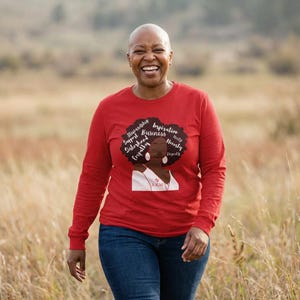 Op de afbeelding: Een vrouw draagt een rood shirt met lange mouwen met een afbeelding van een vrouwen gezicht en de woorden "Stewardship, Support, Sisterhood, Empathy, Business, Inspiration, Unity, Honesty, Dignity". Ze lacht en loopt in een veld.