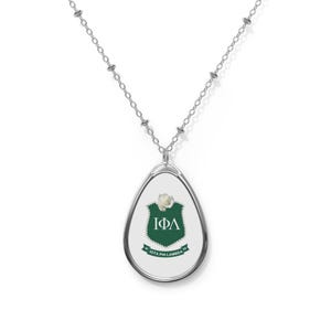 Op de afbeelding: Een zilveren ketting met een traanvormige hanger met een witte parel en een groen schild met de letters "IΦΛ" en de tekst "IOTA PHI LAMBDA" in wit.