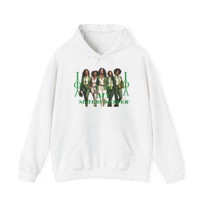 Puede incluir: Sudadera con capucha blanca con bolsillo delantero. El gráfico en la parte delantera presenta un grupo de mujeres con atuendos verdes y blancos, con el texto "Sisters Keeper" en verde. El diseño incluye letras griegas.