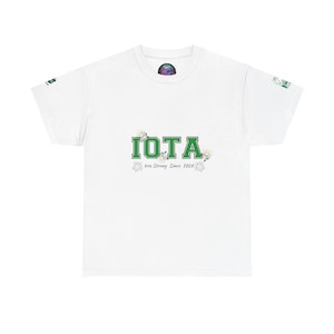 Puede incluir: Camiseta blanca con la palabra "IOTA" en letras verdes. Debajo está el texto "Iota Strong Since 1929". La camiseta tiene cuello redondo y mangas cortas. Un diseño colorido es visible en la parte superior de la espalda.