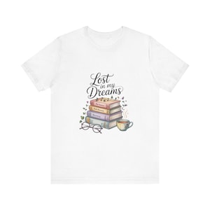 May include: White t-shirt featuring a watercolor illustration of a stack of books, a teacup, and eyeglasses. The text "Lost in my Dreams" is written above the books in a script font. The design includes green vines and small flowers.