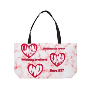 Op de afbeelding: Witte draagtas met zwarte handvatten. De tas heeft een rood marmerpatroon en rode hartvormige afbeeldingen. De harten bevatten de woorden "LAMBDA", "KAPPA" en "MU". Tekst bevat "Sisterhood is Power" en "Since 1937".