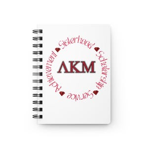 Op de afbeelding: Een wit spiraalgebonden notitieboek met de letters "AKM" in een rood en grijs ontwerp. De letters zijn omcirkeld door de woorden "Achievement, Sisterhood, Scholarship, Service" in rood script, met rode diamantaccenten. Het notitieboek is een briefpapierartikel.