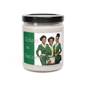 Könnte beinhalten: Eine weiße Kerze in einem Glas mit schwarzem Deckel. Das Etikett zeigt den Text "Sisters in Success" und eine Illustration von drei Frauen in grünen Anzügen und gelben Krawatten. Die Kerze ist ein dekorativer Artikel.
