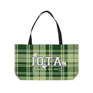 Könnte beinhalten: Eine grüne karierte Tragetasche mit schwarzen Griffen. Die Tasche zeigt das Wort "IOTA" in weißen Blockbuchstaben, mit dem Text "Iota Strong Since 1929" darunter. Weiße Rosenillustrationen sind ebenfalls vorhanden.