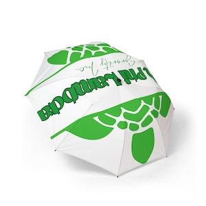 Può includere: Ombrello bianco con dettagli e testo verdi. L'ombrello presenta un motivo verde ripetuto e le parole "Phi Lambda" e "Sorority Inc." in lettere verdi. L'ombrello è aperto e angolato.