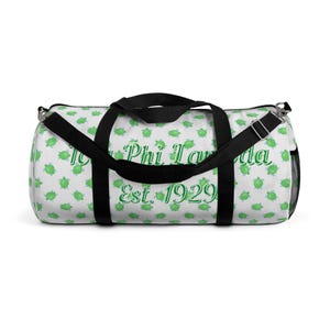 May include: White duffel bag featuring a pattern of green turtles. It has black handles and a shoulder strap. The text "Iota Phi Lambda" and "Est. 1929" are printed in green on the bag.