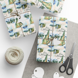 May include: Several wrapped gifts with New Hampshire map-themed wrapping paper. The paper features watercolor illustrations of the state's landmarks and the text "New Hampshire". Each package is tied with twine and a white heart-shaped tag. A ball of twine, scissors, and blank tags are also visible.