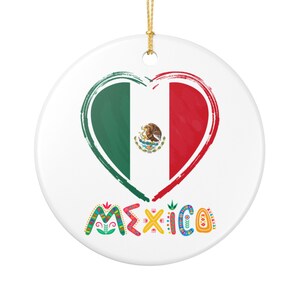 Op de afbeelding: Een witte keramische ornament met een hartvormig ontwerp in de kleuren van de Mexicaanse vlag: groen, wit en rood. Het woord "MEXICO" is in kleurrijke, decoratieve letters onder het hart geschreven. Een gouden touwtje is bevestigd om op te hangen.