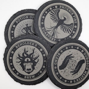 Puede incluir: Cuatro posavasos de pizarra negra con un diseño circular. Cada posavasos presenta un gráfico y un texto diferentes. Los posavasos representan un fénix, una poción, un beholder y un par de manos sosteniendo un dado. El texto en los posavasos dice "Phoenix Fire Roastery", "Potion of Brew", "Beholder's Brew" y "Dungeon Master's Blend".