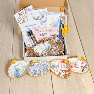May include: A craft kit for making decorative shell dishes. The kit includes four unfinished white ceramic shells, decoupage paper with bird designs, paint, brushes, and scissors. The kit also includes instructions and a card that says "Cass + Pearl".