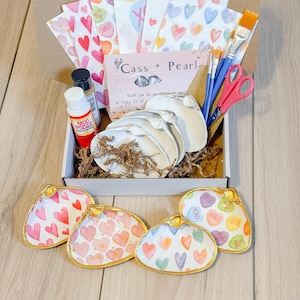 May include: A craft kit for making heart-shaped trinket dishes. The kit includes four unfinished white ceramic dishes, paint, brushes, and decorative paper with hearts and the words "Be Mine" and "Sweet".