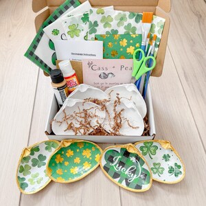 May include: A craft kit for making decorative shell dishes. The kit includes four white ceramic shells, green and gold patterned paper napkins, paintbrushes, glue, and a card that says "Cass + Pea".