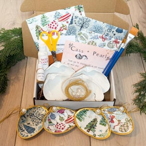 May include: A craft kit in an open cardboard box, featuring seashell ornaments with gold trim and festive designs. Includes Mod Podge, paintbrushes, scissors, and decorative paper. The kit also contains the text "Cass + Pearl".