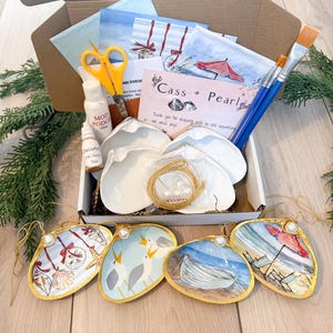 May include: An open cardboard box filled with craft supplies. Includes white scallop shells, paintbrushes, paint, glue, scissors, and painted shell ornaments with gold trim. The ornaments feature beach scenes and holiday designs.