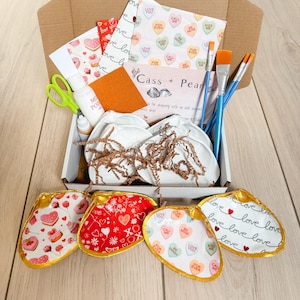 Könnte beinhalten: Ein Bastelset in einem Karton mit herzförmigen Schalen mit goldenen Rändern. Das Set enthält Pinsel, Scheren, Mod Podge und Dekopapier mit Herzen und dem Text "Love You". Die Schalen sind mit Herzen und den Worten "Love", "Be Mine" und "Kiss Me" verziert.