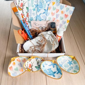 May include: A craft kit containing seashell-shaped dishes, paintbrushes, scissors, and decorative napkins. The dishes display colourful designs of butterflies, hearts, and marine life. The kit includes paint and glue.