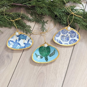 May include: Three seashell ornaments with gold trim and pearl accents. The shells feature blue and white patterns, including turtles and floral designs. Each ornament hangs from a gold string, perfect for coastal or nautical decor.
