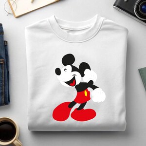 May include: White crewneck sweatshirt featuring a smiling Mickey Mouse graphic. The cartoon character is depicted in his classic outfit: red shorts, yellow buttons, black body, and white gloves. The sweatshirt is made of a soft, comfortable material.