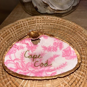 以下が含まれることがあります： ピンクの花柄と金色の縁取りが施された白い貝殻。貝殻には金色の文字で「ケープコッド」と書かれています。