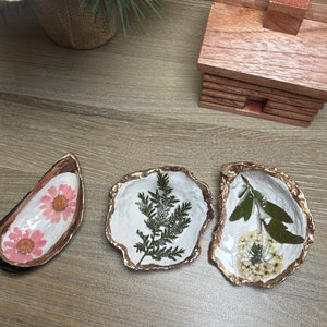 May include: Three small white oyster shells with gold trim, each filled with a different pressed flower or plant. The shells are arranged on a light brown wooden surface.