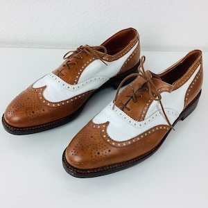 May include: A pair of two-tone leather Oxford shoes. The shoes feature a brown toe and heel, white center panel, and brown laces. The shoes have decorative perforations and are set against a white background.