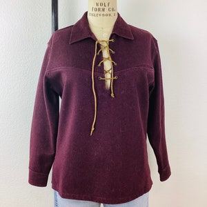 May include: A burgundy long-sleeve shirt with a collared neckline and a lace-up front. The shirt is displayed on a mannequin. The lace is a light brown color. The shirt has a classic, vintage style.