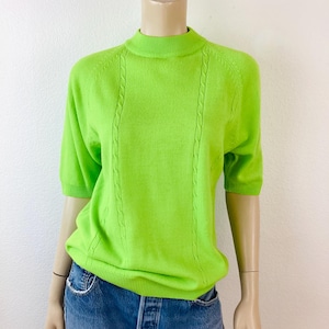 May include: A bright green short-sleeve sweater with a mock neck. The sweater features a cable knit design down the front and is displayed on a mannequin. The sweater is paired with blue jeans.