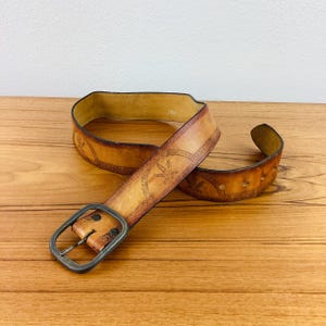 May include: A light brown leather belt with a rectangular, dark metal buckle. The belt features embossed floral and starburst designs. The belt is lying on a wooden surface.