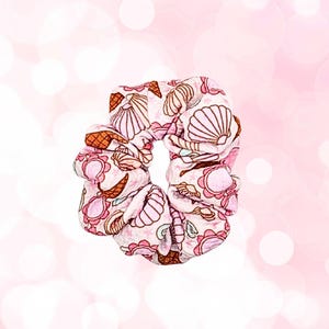 May include: A pink scrunchie with a seashell pattern. The fabric features light pink, brown, and white seashells, ice cream cones, and floral designs. The scrunchie is a hair accessory.