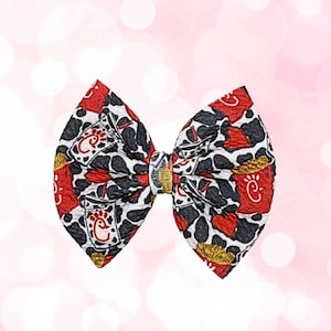 Puede incluir: Un lazo para el pelo de tela con un patrón repetido de elementos rojos, negros y dorados sobre un fondo blanco. El lazo presenta un nudo central con detalles dorados. El patrón incluye un logotipo estilizado.